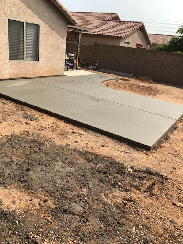 Newly poured concrete patio in a backyard with dirt, a house, and a fence.