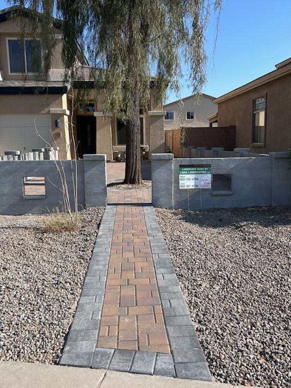Brick pathway leading to a house, framed by low grey walls and a tree. Brown and grey brickwork with gravel ground.