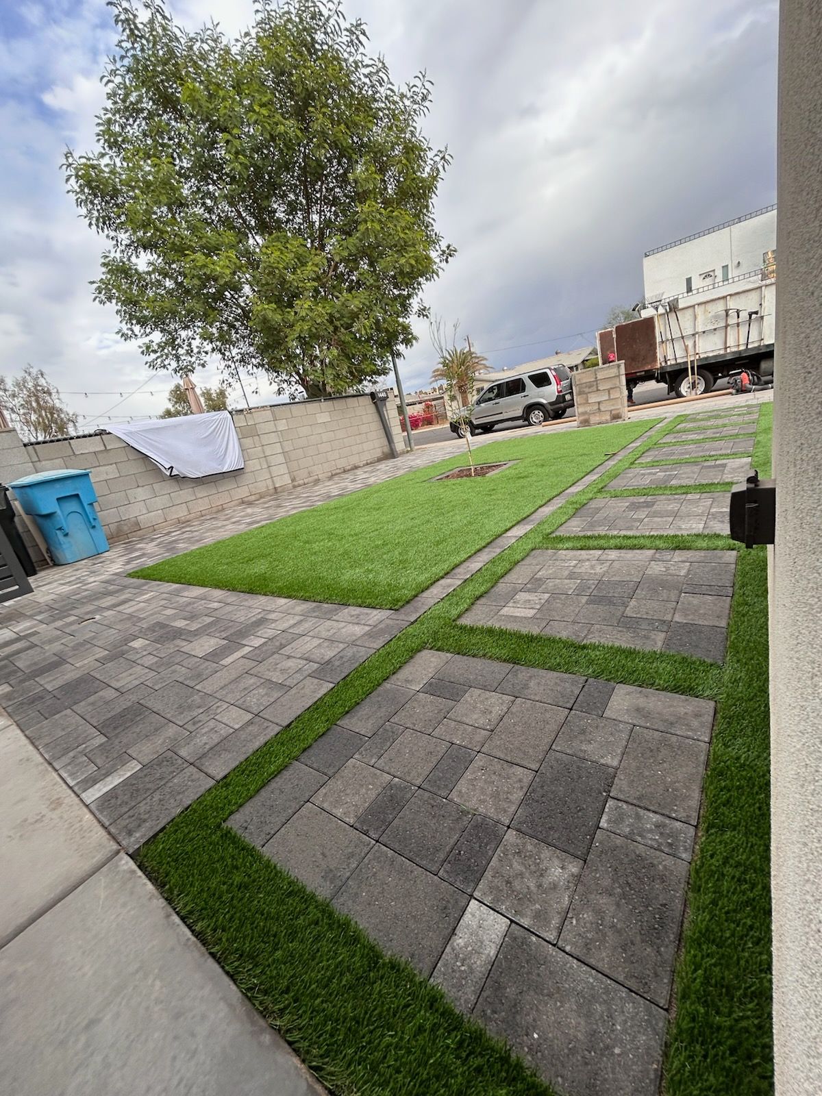 Paver pathway with green strips, leading to a lawn with a tree.