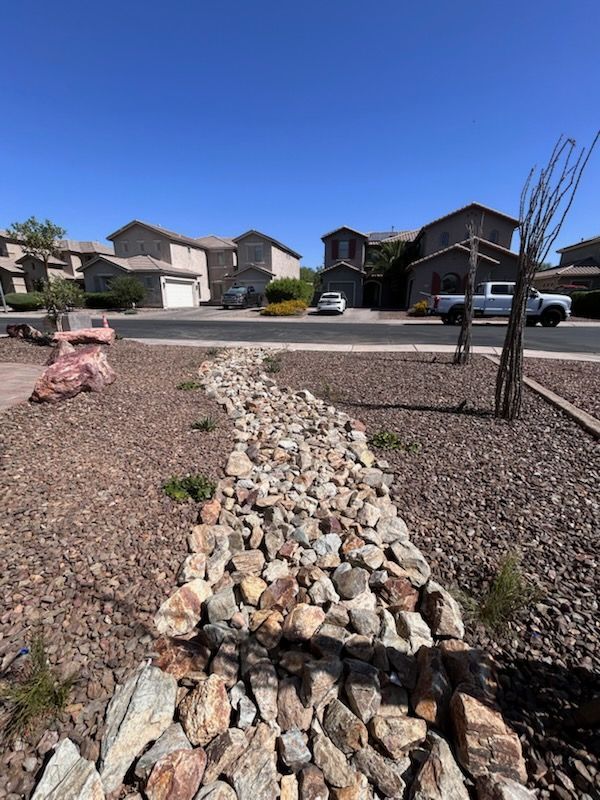 Rock-lined drainage swale leading toward suburban homes under a clear blue sky