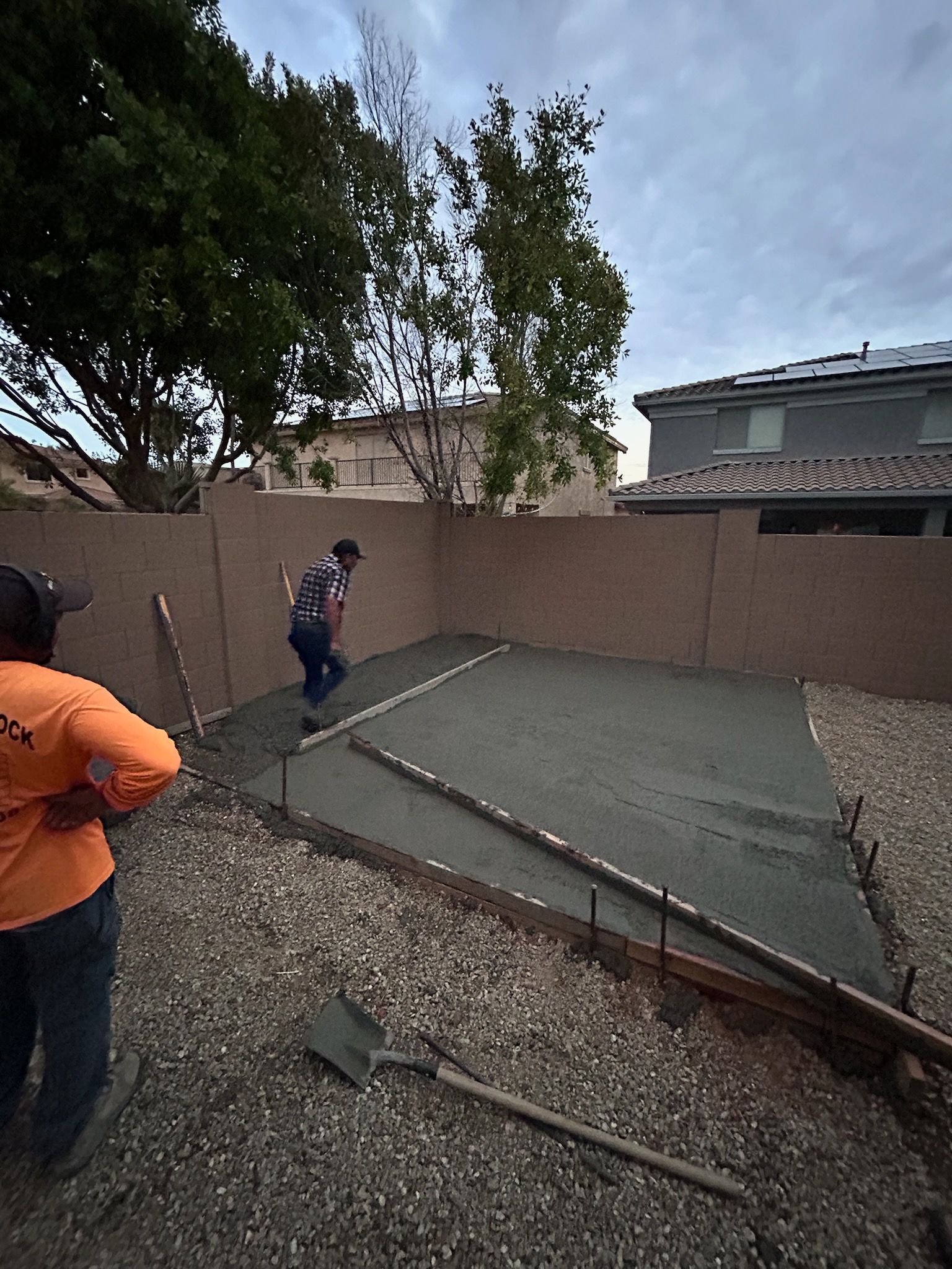 Workers leveling wet concrete in a backyard construction area with gravel and a fence.