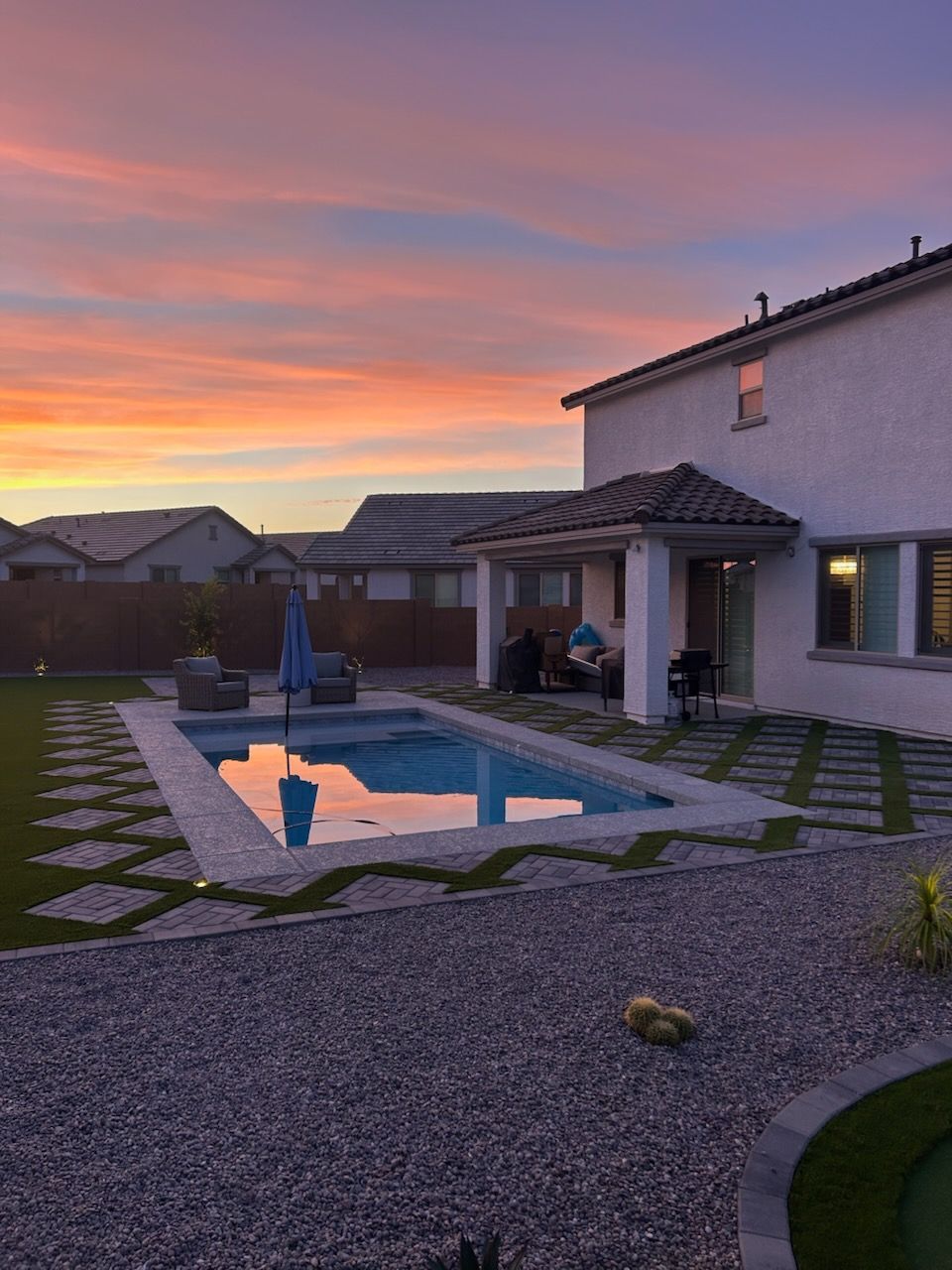 Backyard pool beside a white house at sunset, with pink-orange sky and gravel patio