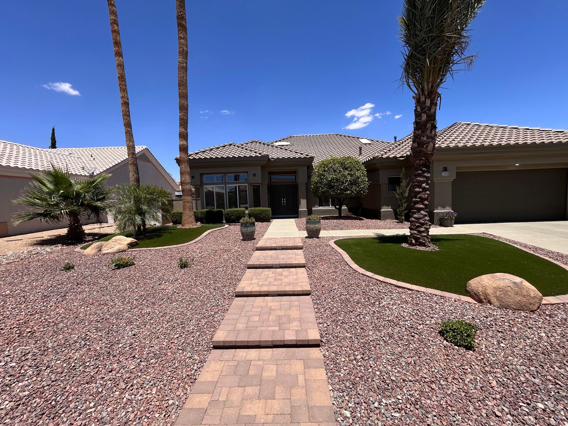 Front yard desert landscaping with a brick walkway leading to a single-story house under a blue sky