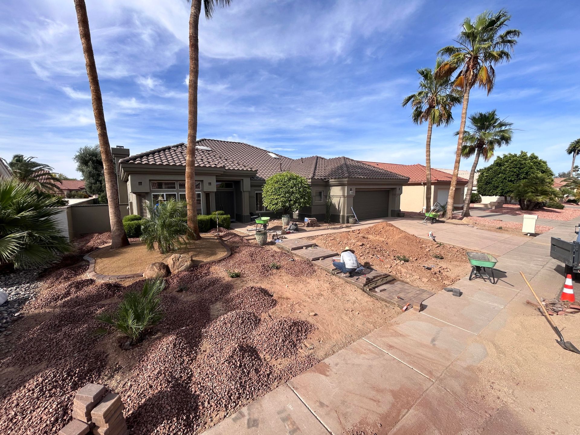 Suburban house under construction with dirt piles, palm trees, and a partly paved driveway