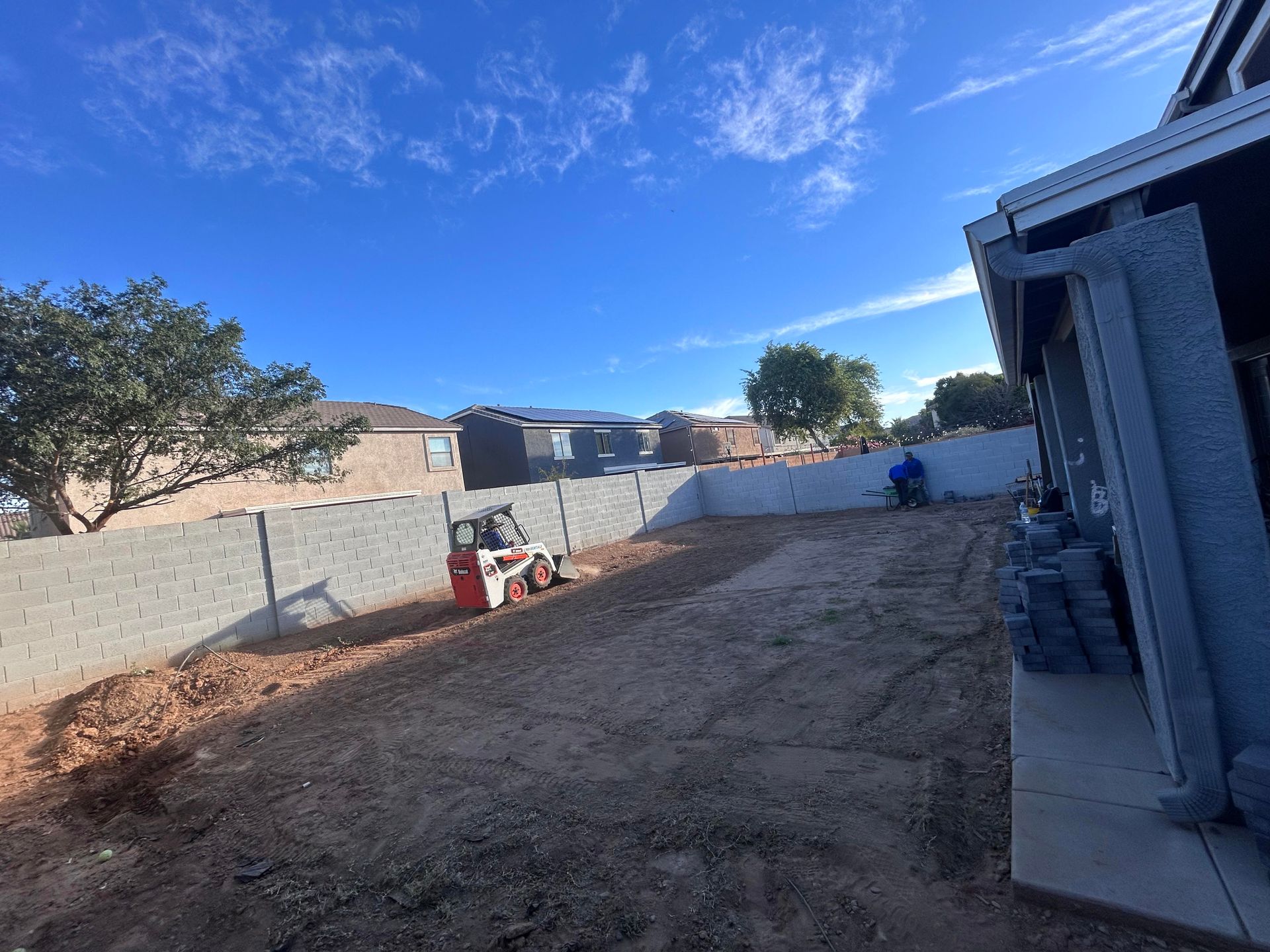 Backyard with dirt ground, gray walls, a tree, and a bright blue sky