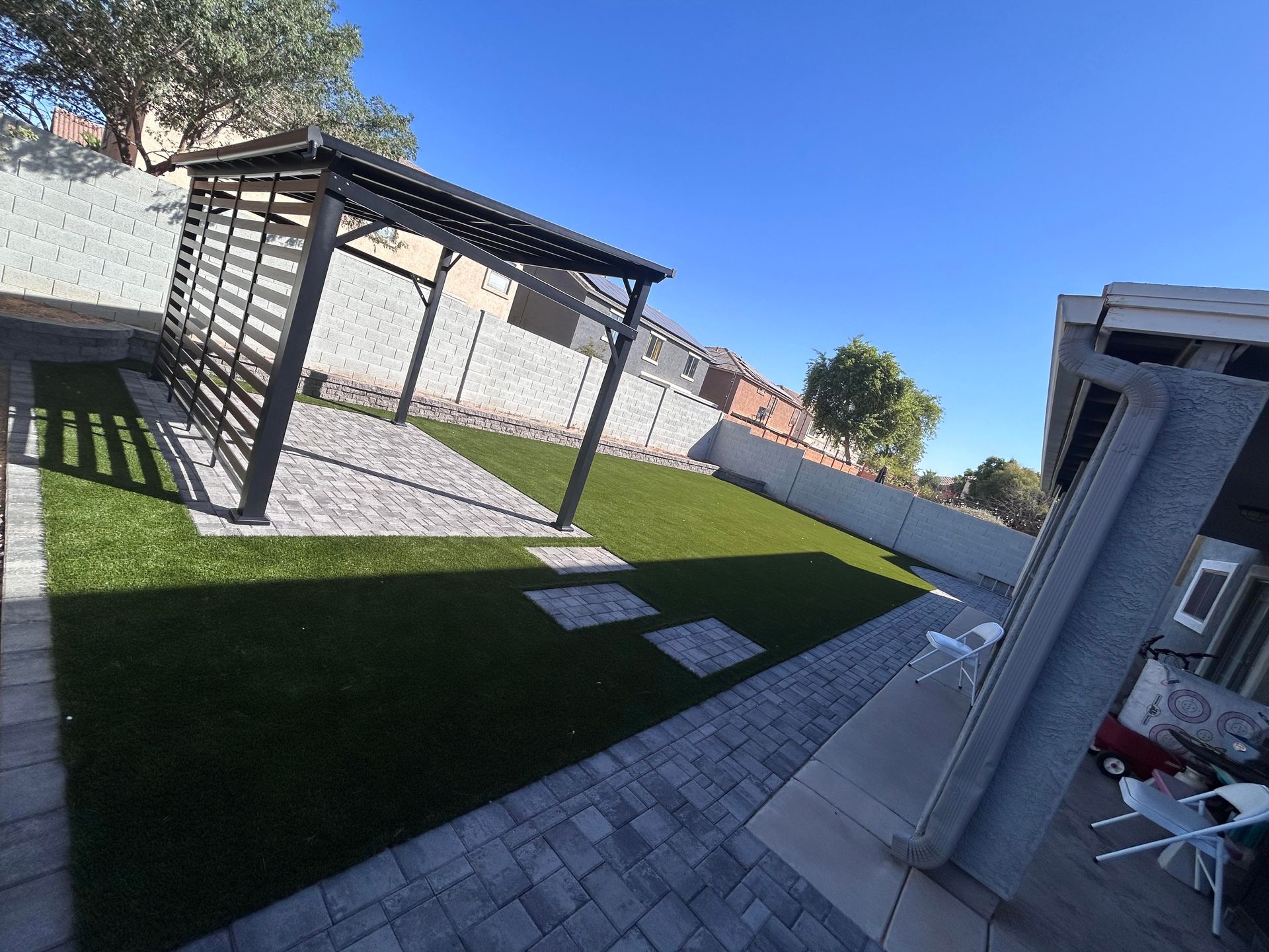 Backyard patio with pergola, paved walkway, green lawn, and outdoor seating under a bright blue sky