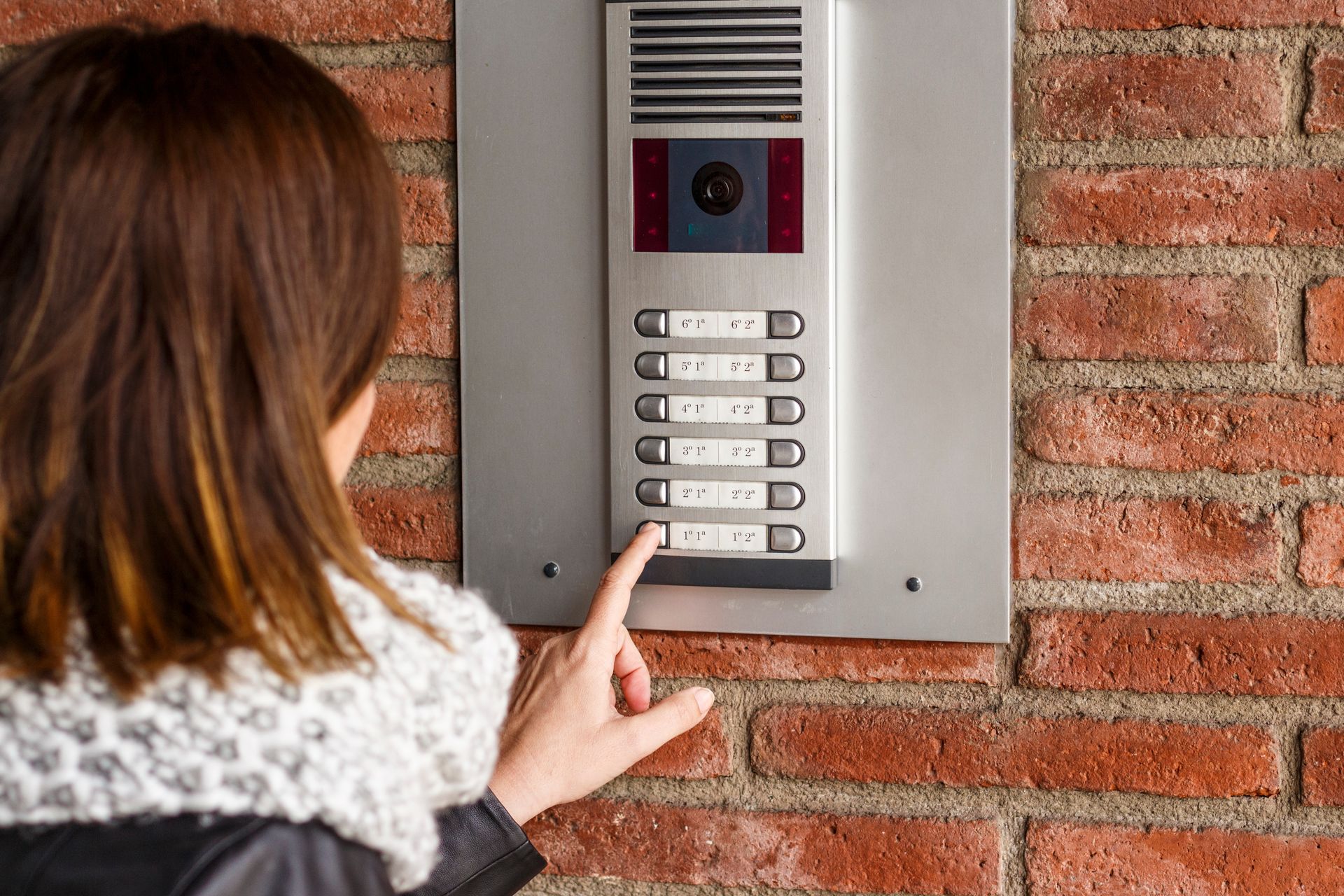 Une personne appuie sur un bouton du panneau d'interphone d'un immeuble d'appartements, fixé sur un mur de briques rouges.