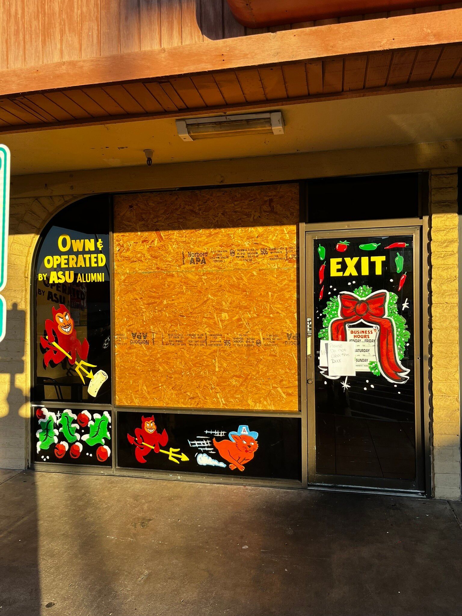 Closed storefront with boarded-up window and an EXIT door decorated with colorful holiday art.