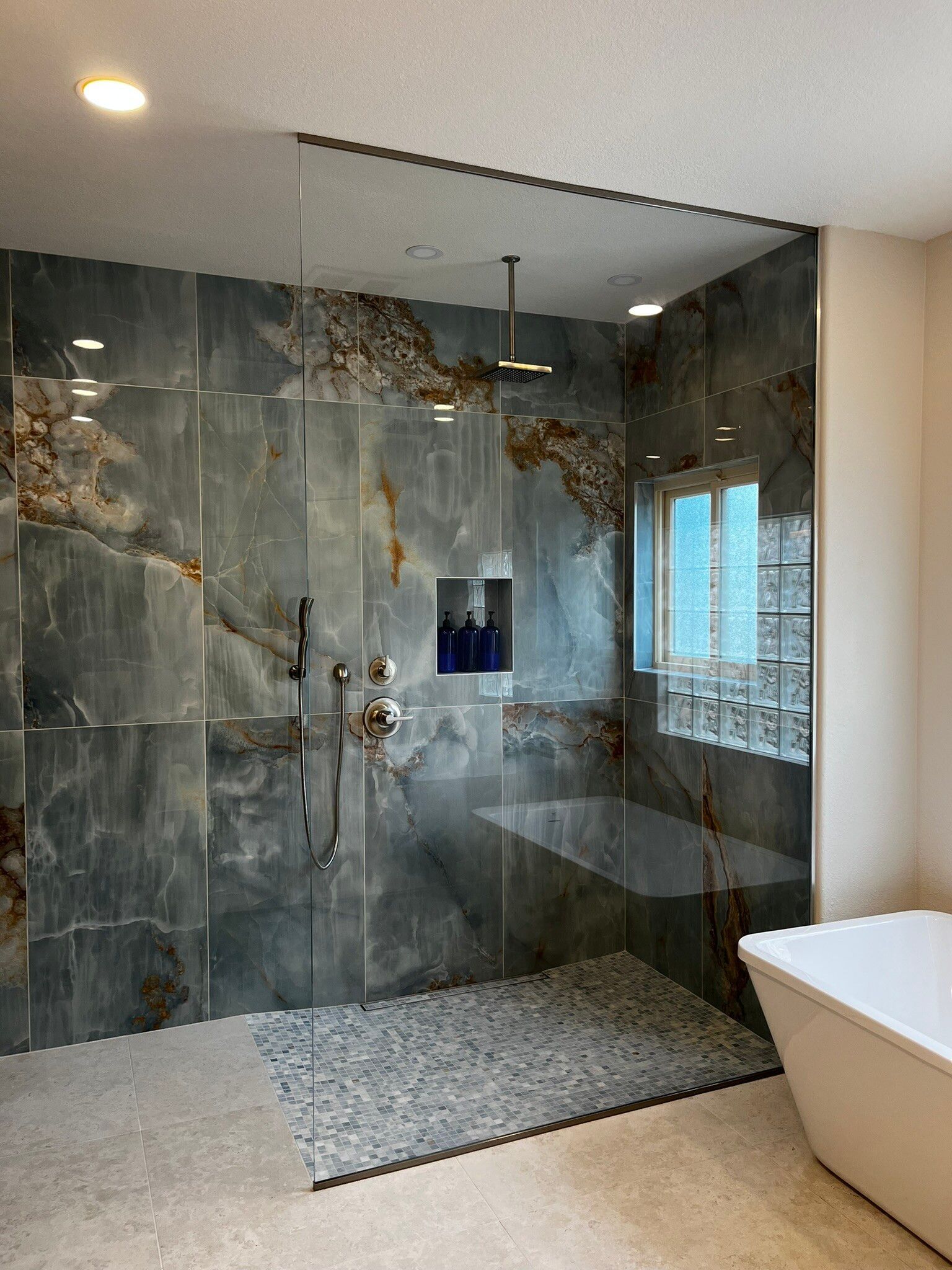 Modern bathroom shower with gray stone walls, glass enclosure, bench, rain shower, and a freestanding tub nearby