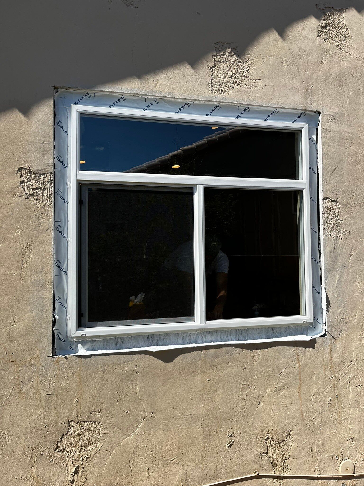 Small white-framed window set in a rough beige wall, with cracked glass and a dark interior behind it.