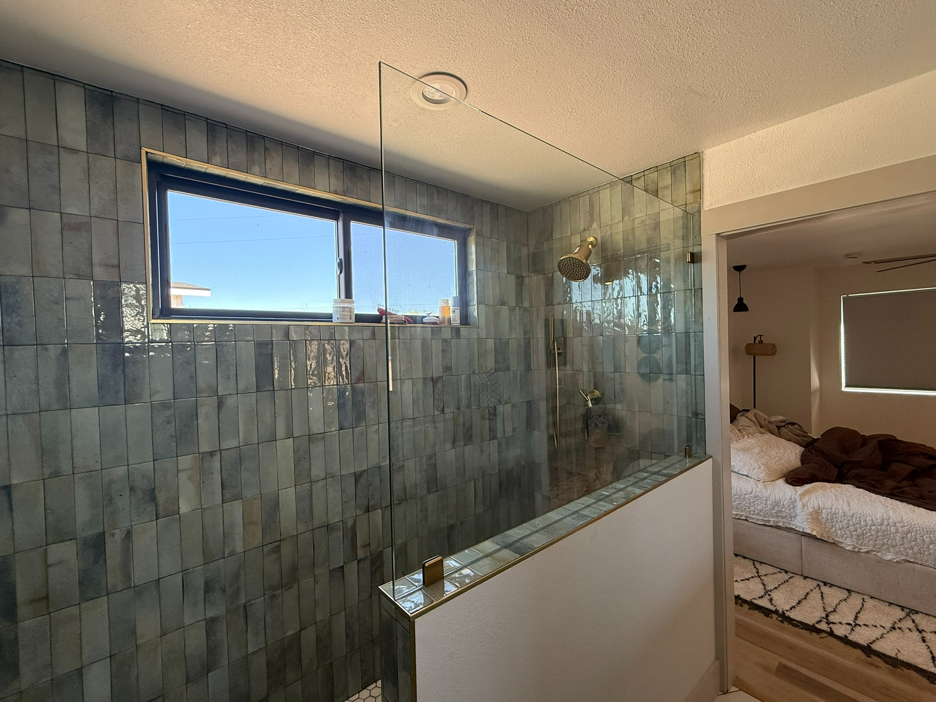 Bathroom with glass shower enclosure, gray tile walls, and a small window beside a bedroom doorway.