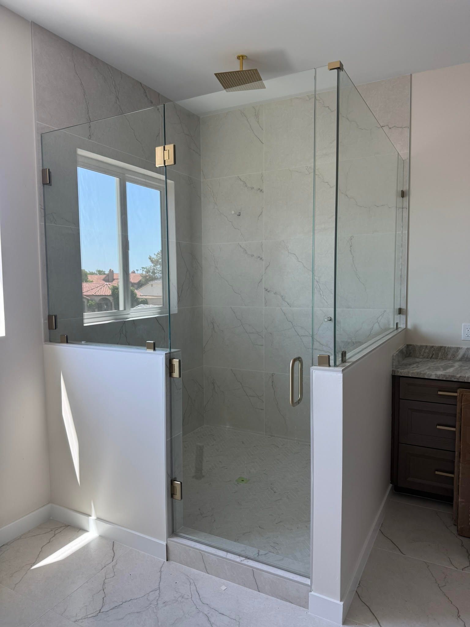Glass shower enclosure in a bathroom with white marble-look walls and an open window nearby