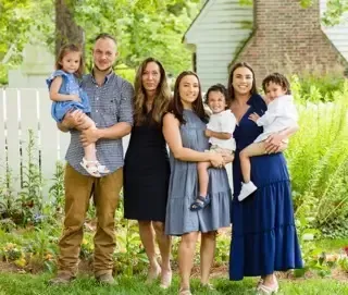 A family of seven stands together in a garden, smiling for a photo in front of a white fence and lush greenery.