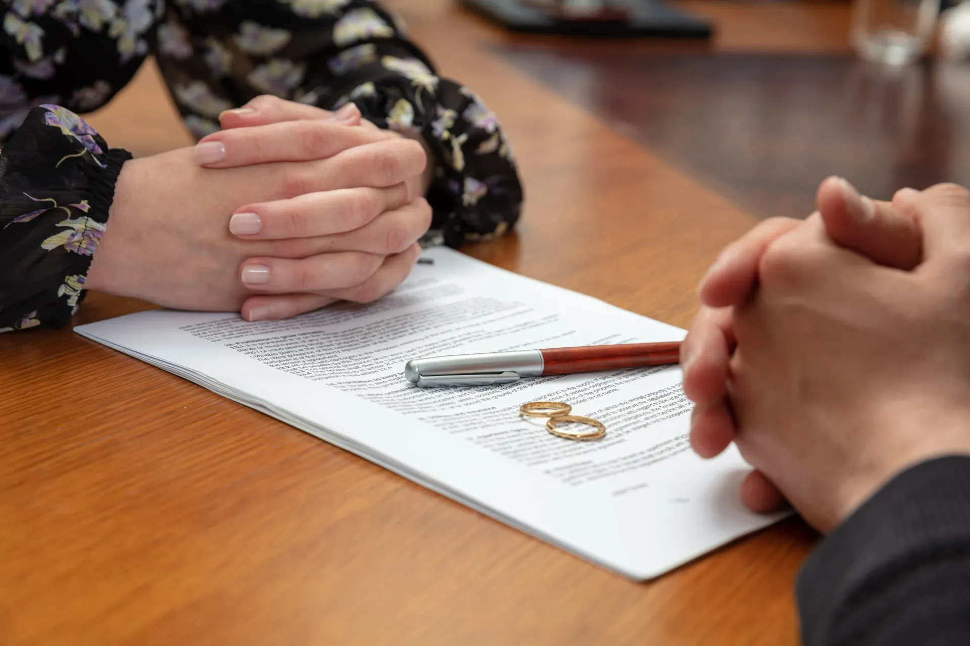 Two sets of hands rest on a table beside a document, a pen, and two gold wedding rings, suggesting a legal separation.