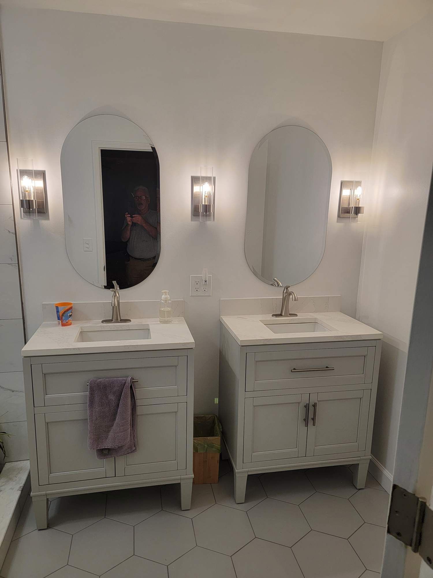 Two white bathroom vanities with oval mirrors and wall sconces in a bright tiled bathroom.