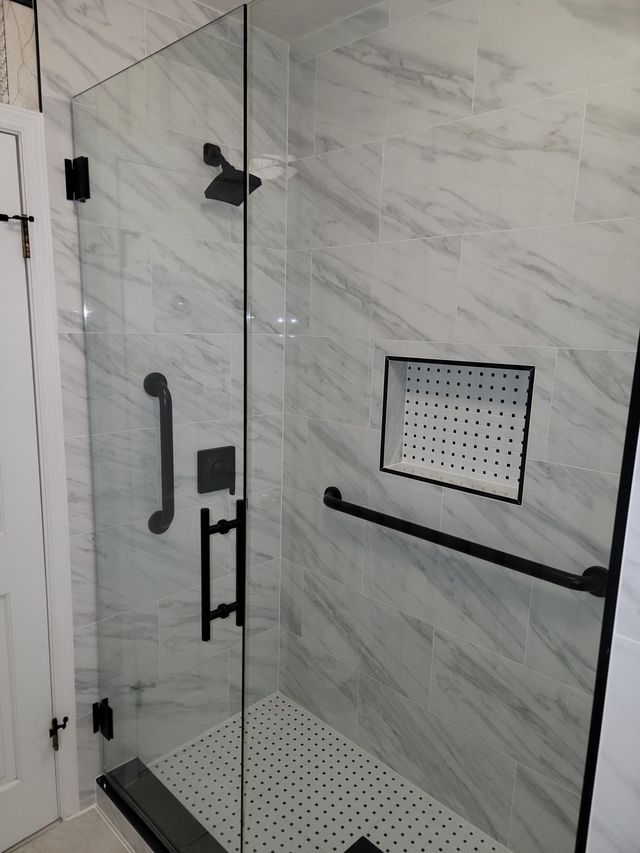 Walk-in shower with glass door, marble tile walls, black grab bars, and a fold-down shower seat