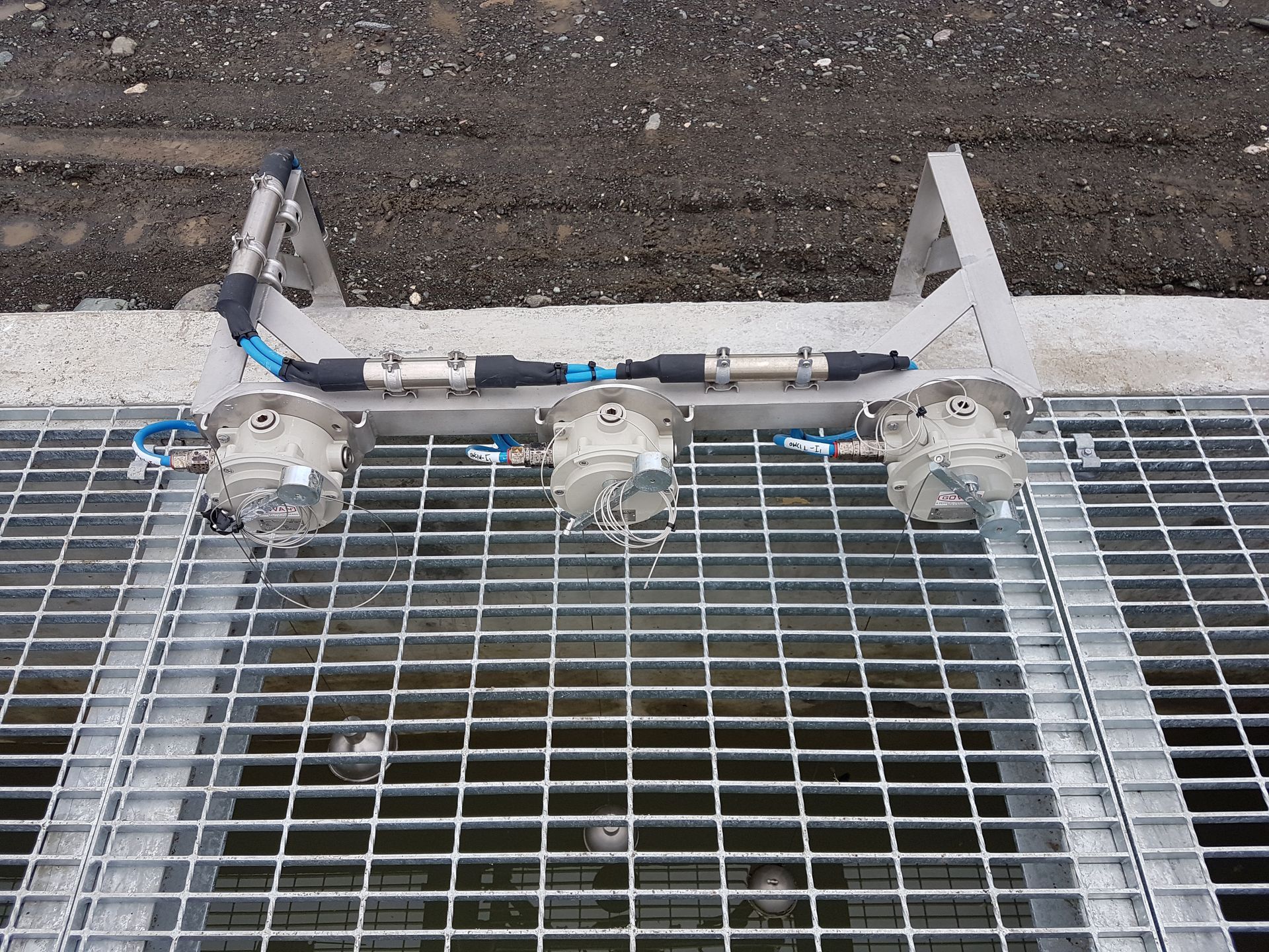 Three gray sensor devices mounted on metal grate, connected by black tubing and metal frame, set outdoors.
