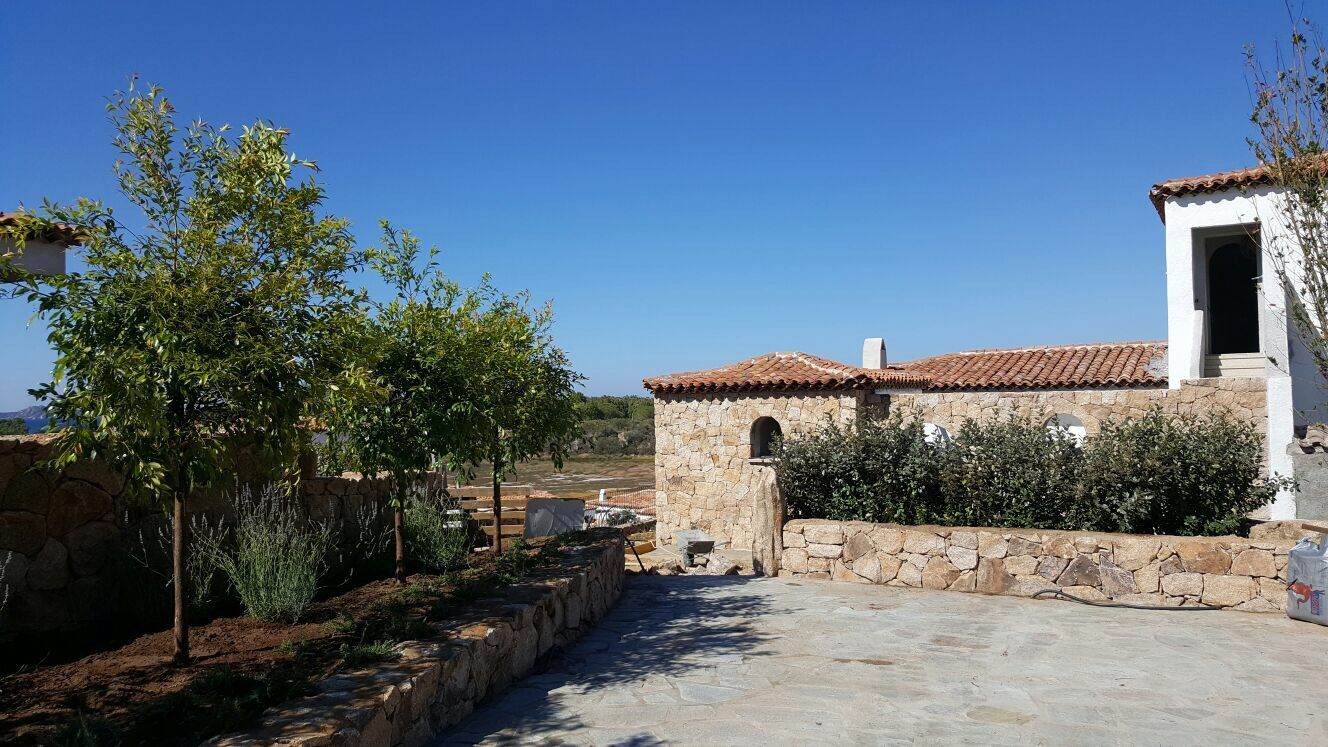 Un edificio in pietra con tetto in tegole è circondato da alberi e cespugli.