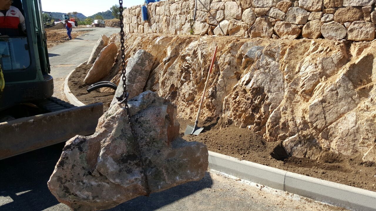 Vor einer Steinmauer wird ein großer Fels von einem Kran angehoben.