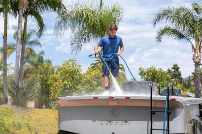 Man power-washing a boat deck in a sunny yard with palm trees and spray mist