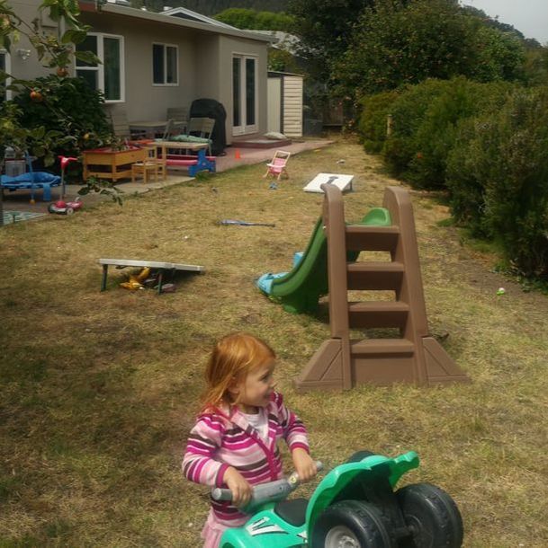 A little girl is riding a green toy motorcycle in a yard.