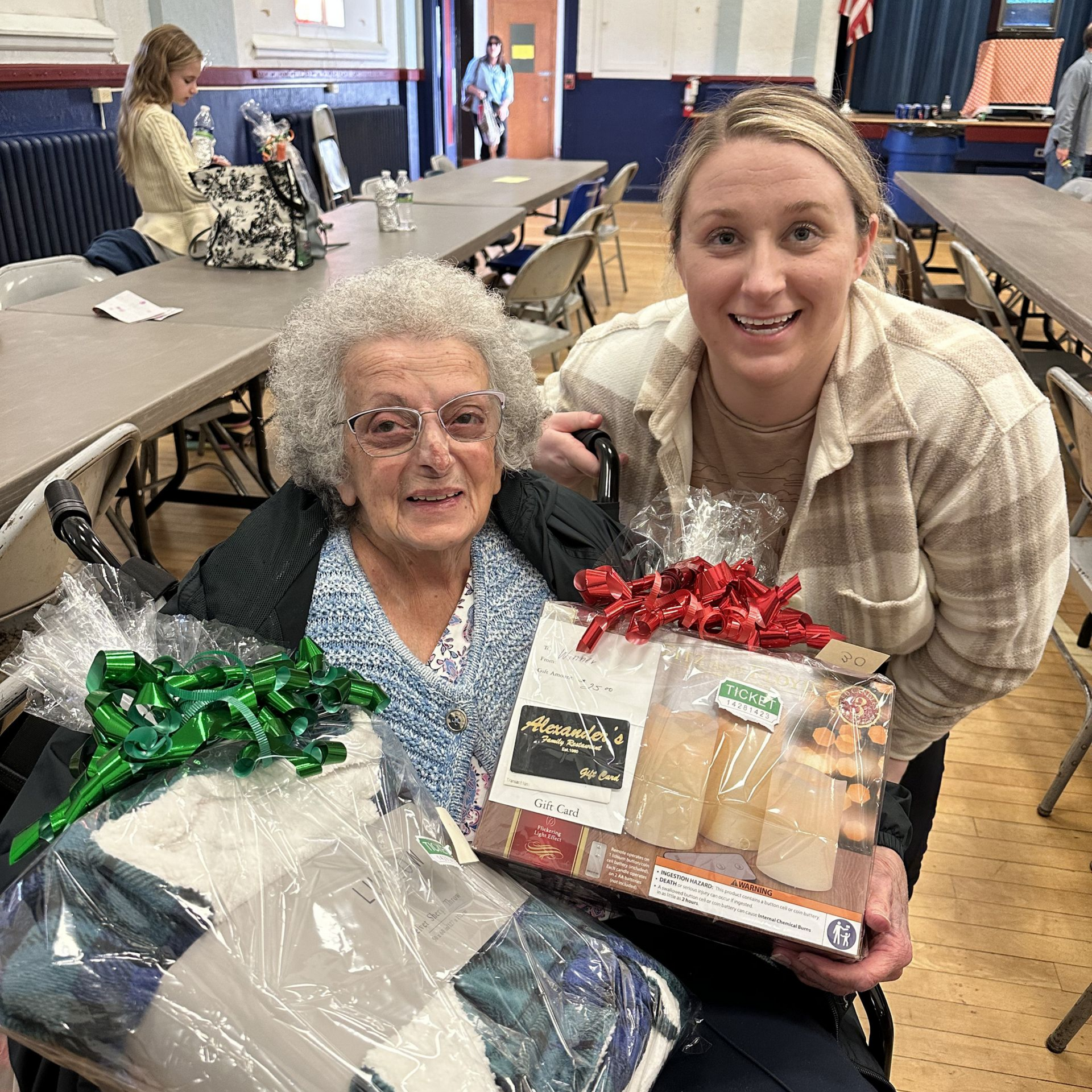 Woman in wheelchair smiles with another woman holding a gift basket and candles in a community hall.