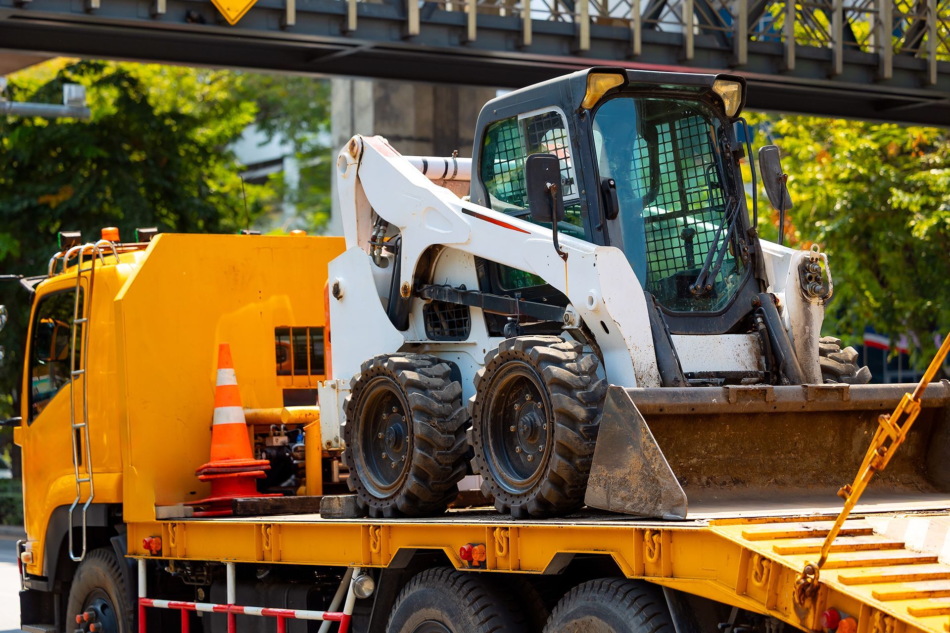 White skid-steer loader on a flatbed truck beside a large yellow machine in an outdoor worksite