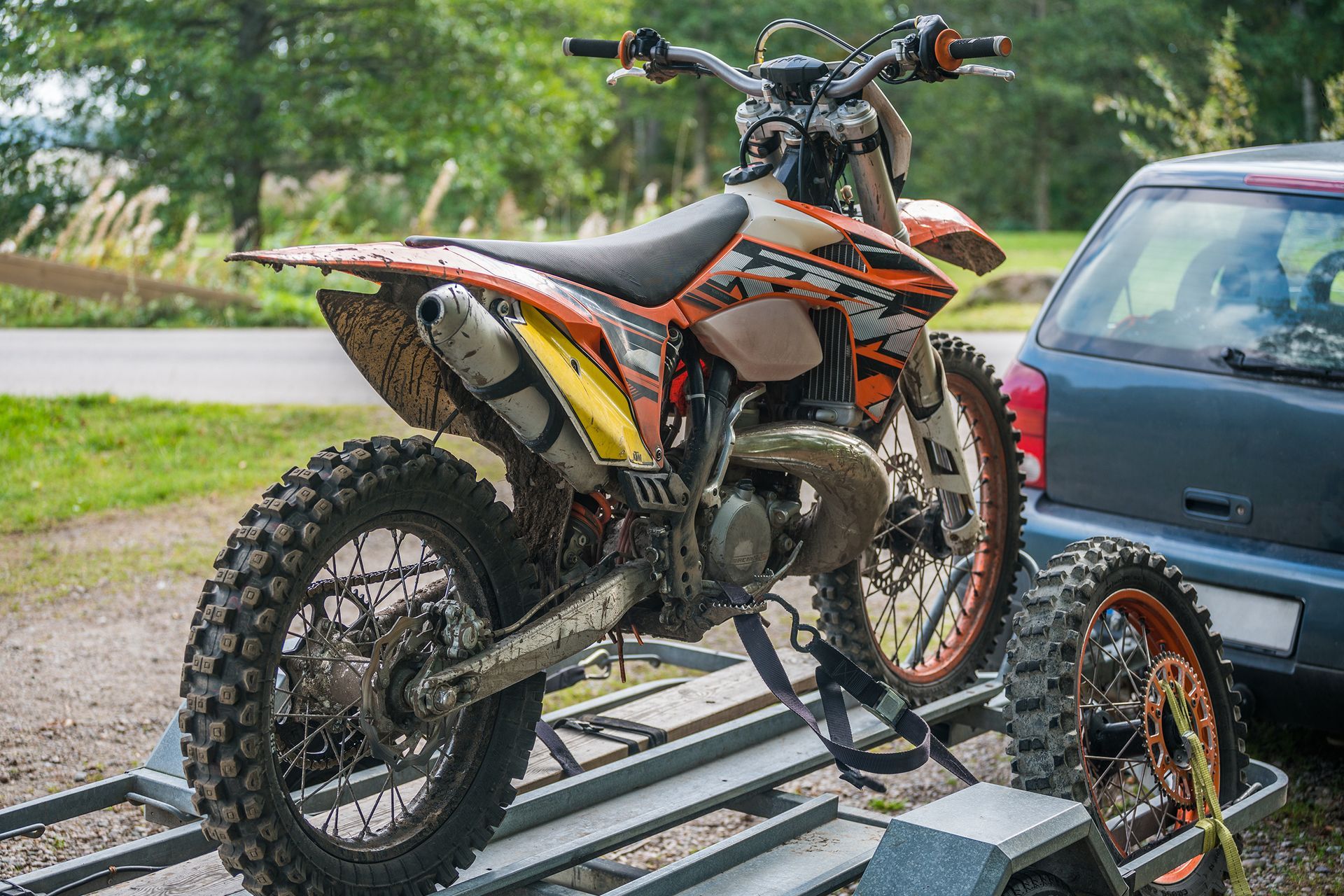 Orange dirt bike strapped to a trailer beside a parked car on a grassy roadside