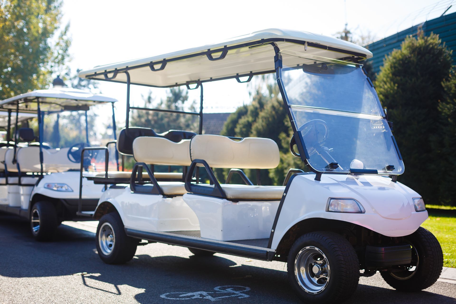 White golf carts lined up outdoors on a sunny street, one with a clear windshield and roof