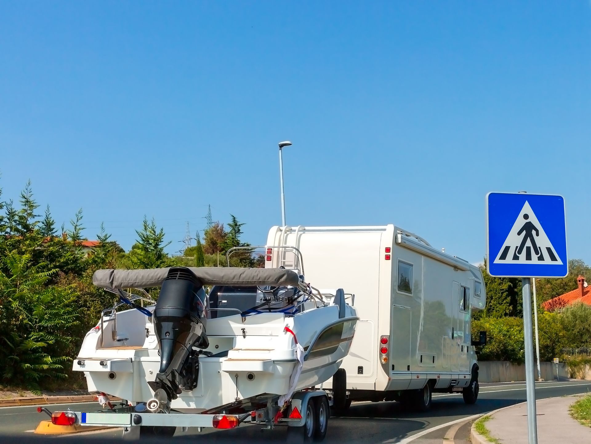 Boat and trailer on roadside near a pedestrian crossing sign, with a white RV behind them.
