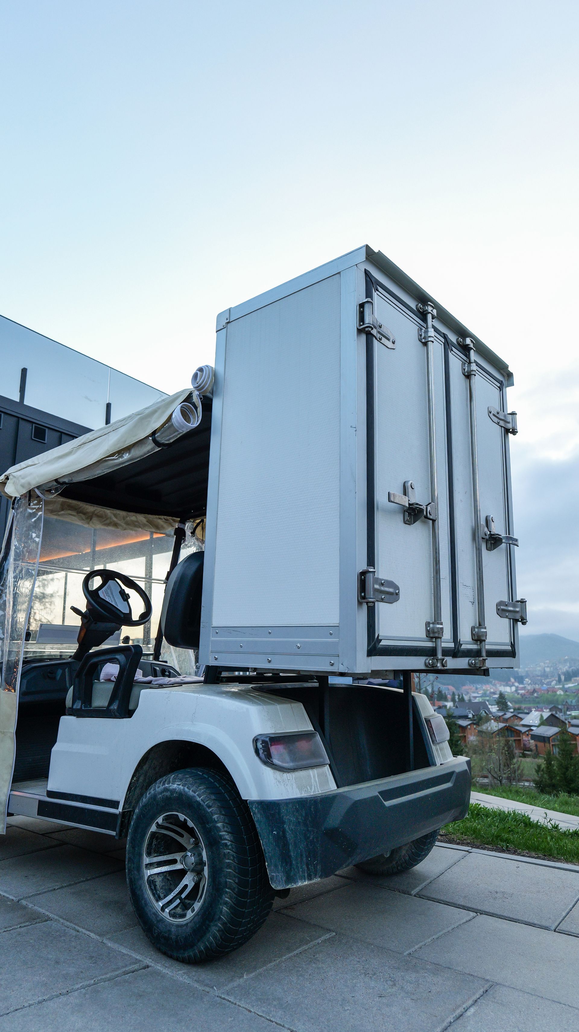 White utility vehicle with a large rear cargo box parked outdoors on a concrete road near buildings.