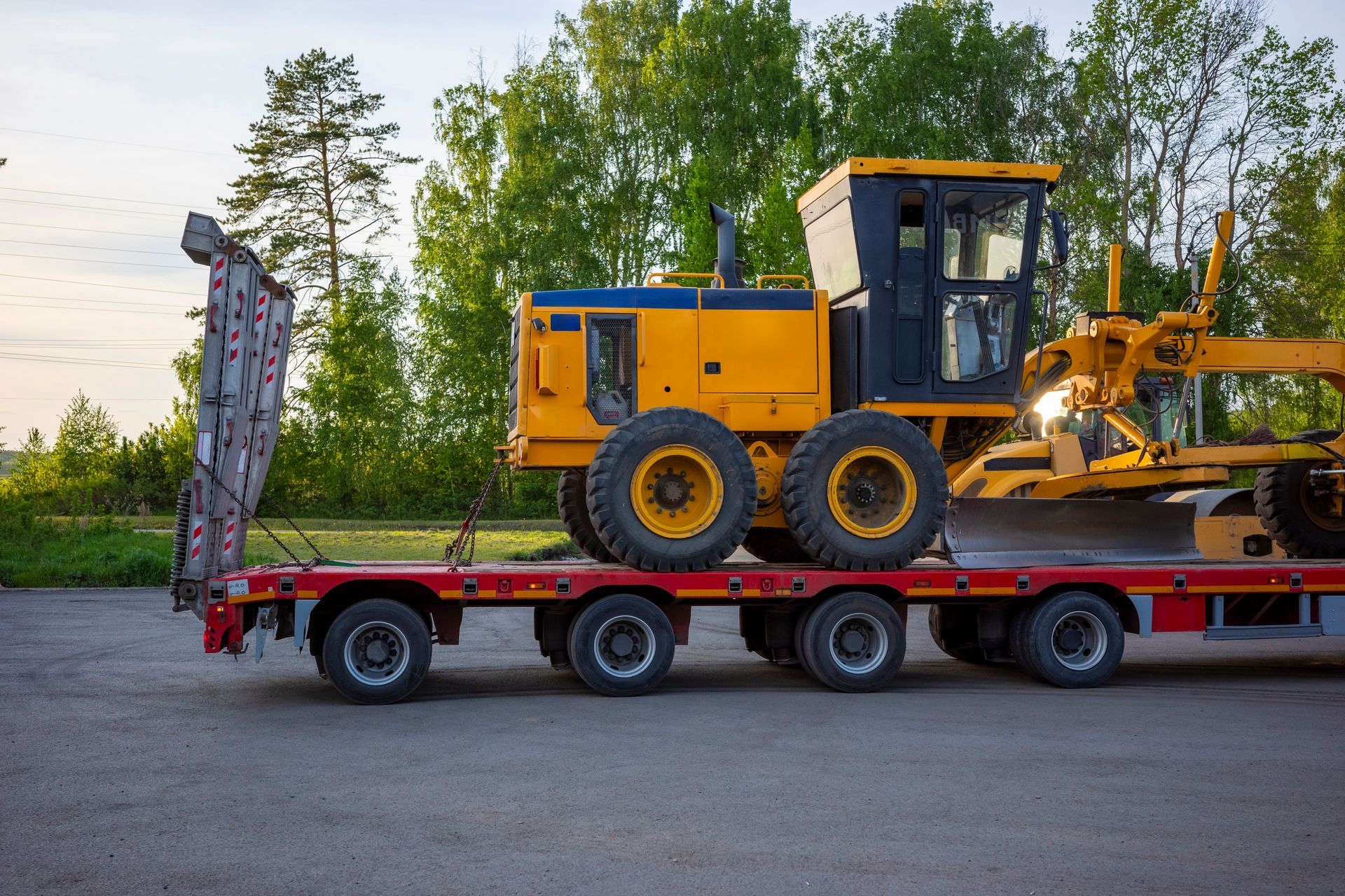 Yellow construction vehicle on a red flatbed trailer in a parking lot with trees in the background