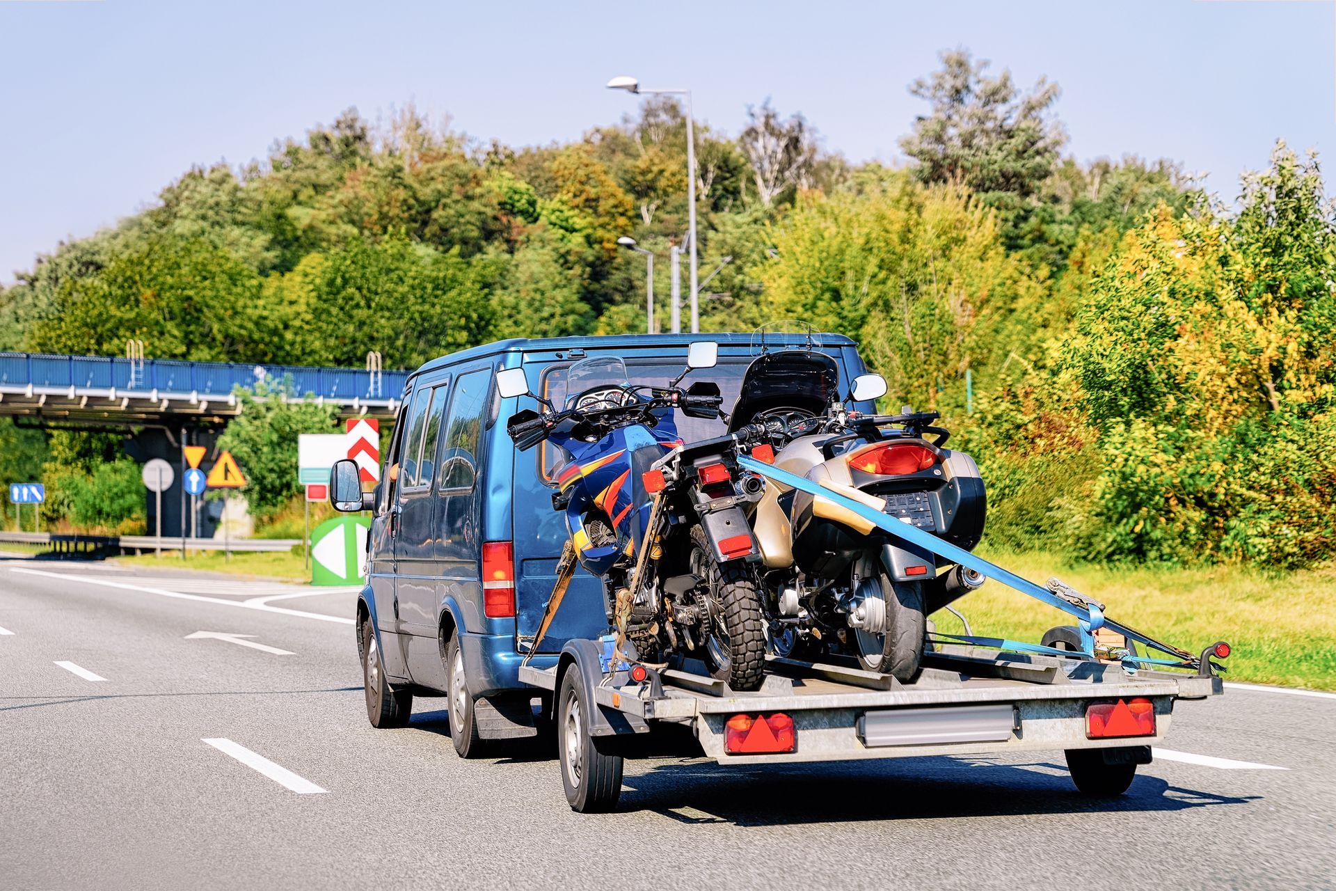 Blue van towing a loaded motorcycle trailer on a highway, with trees and signs in the background