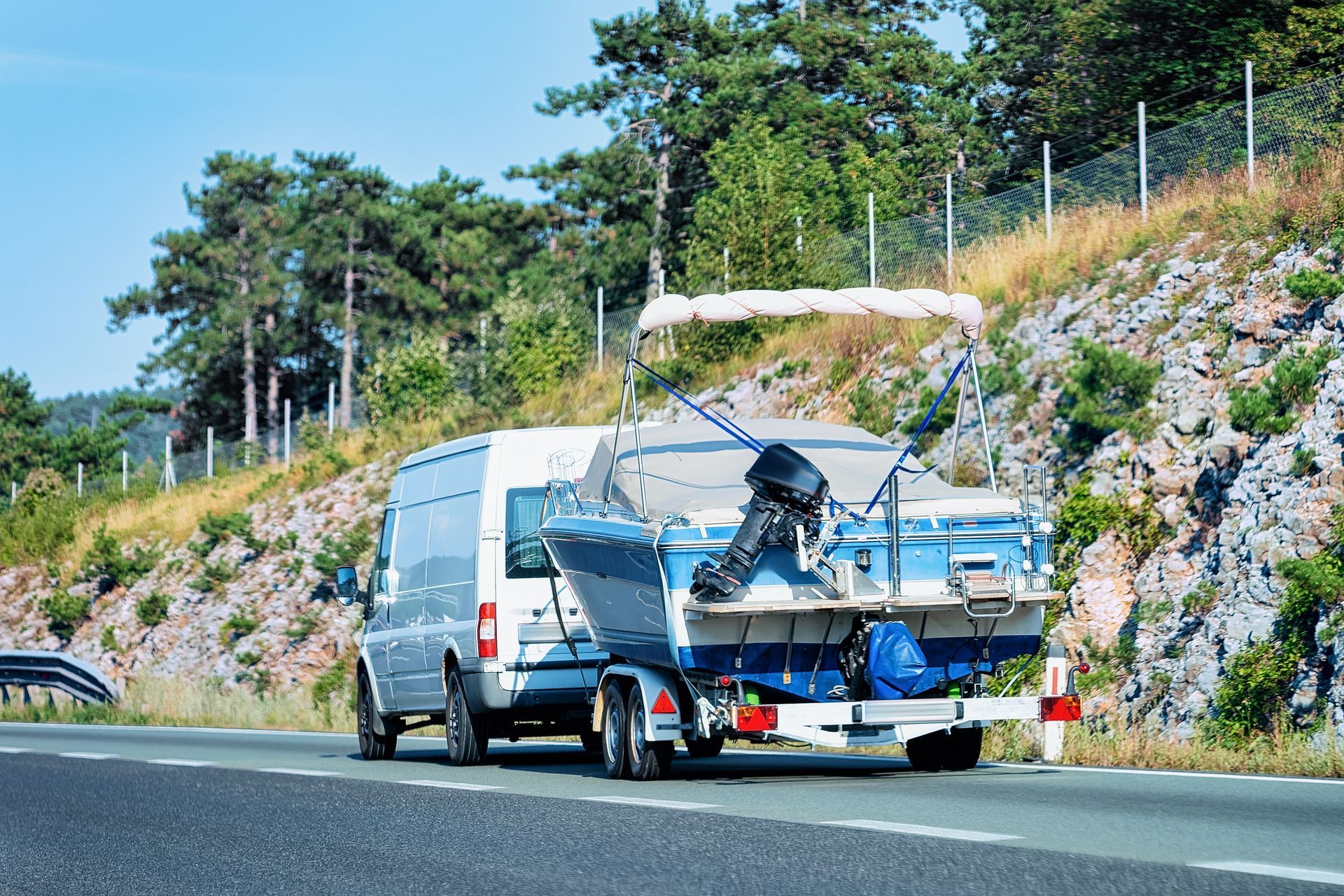 White van towing a blue motorboat on a trailer along a roadside by a rocky hillside