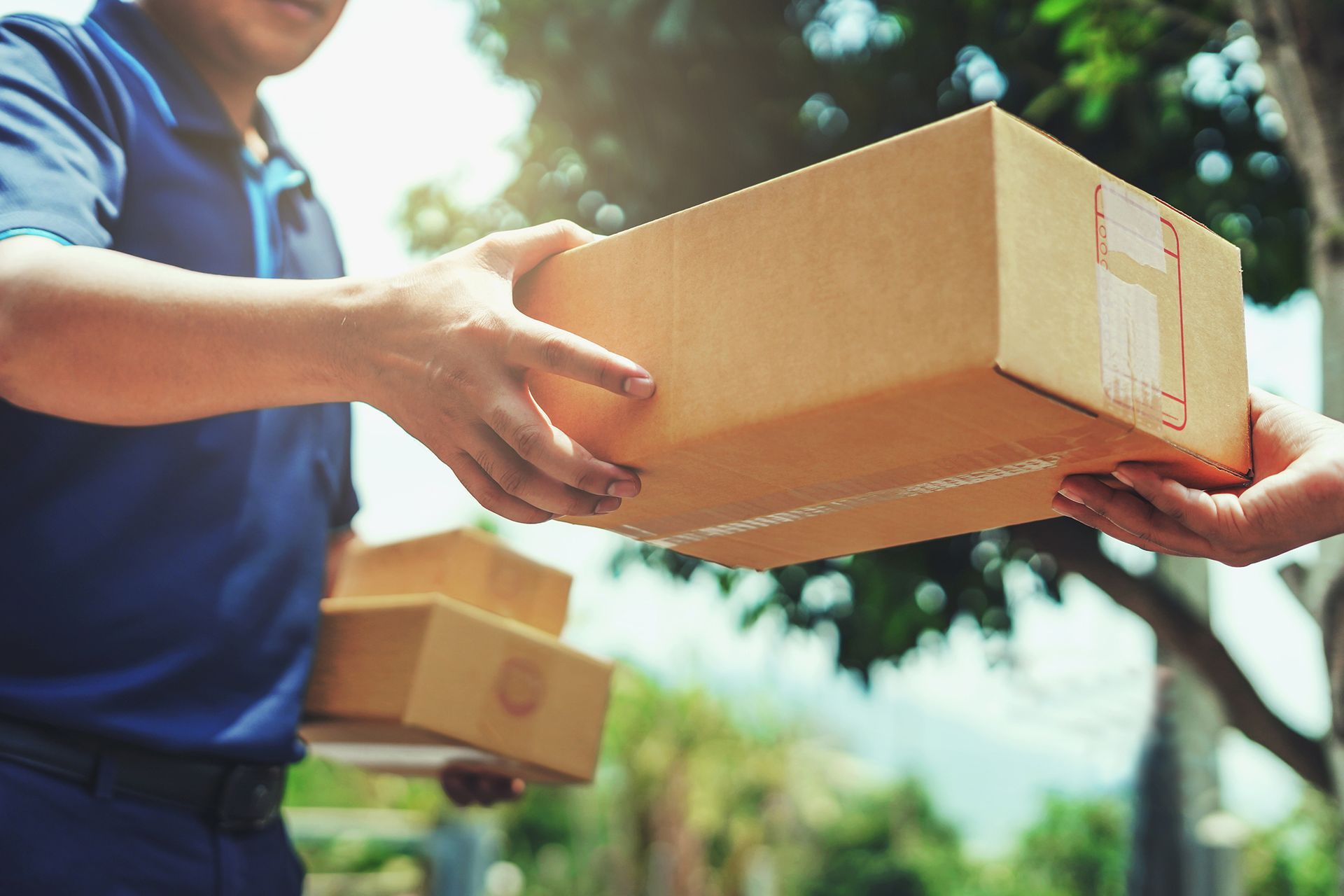 Delivery worker handing a cardboard box to another person outdoors in a sunny setting