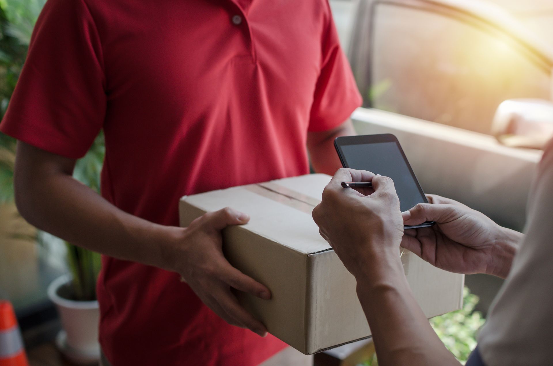 Delivery worker handing a cardboard package to a person at a doorway, with a signature clipboard.