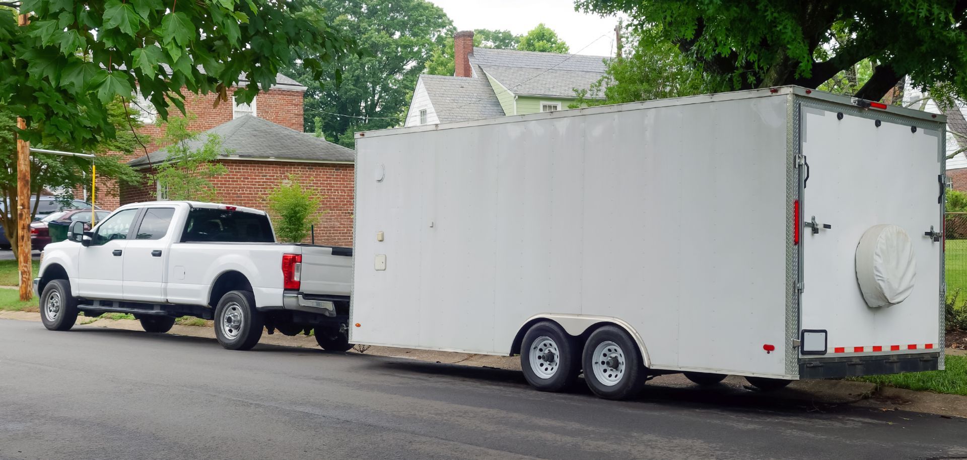 White pickup truck towing a large white enclosed trailer on a suburban street, with trees and houses nearby