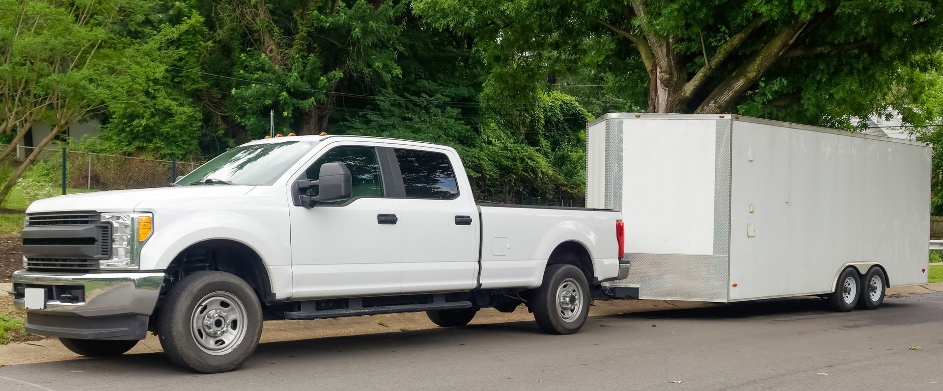 White pickup truck towing a large enclosed white trailer on a tree-lined road