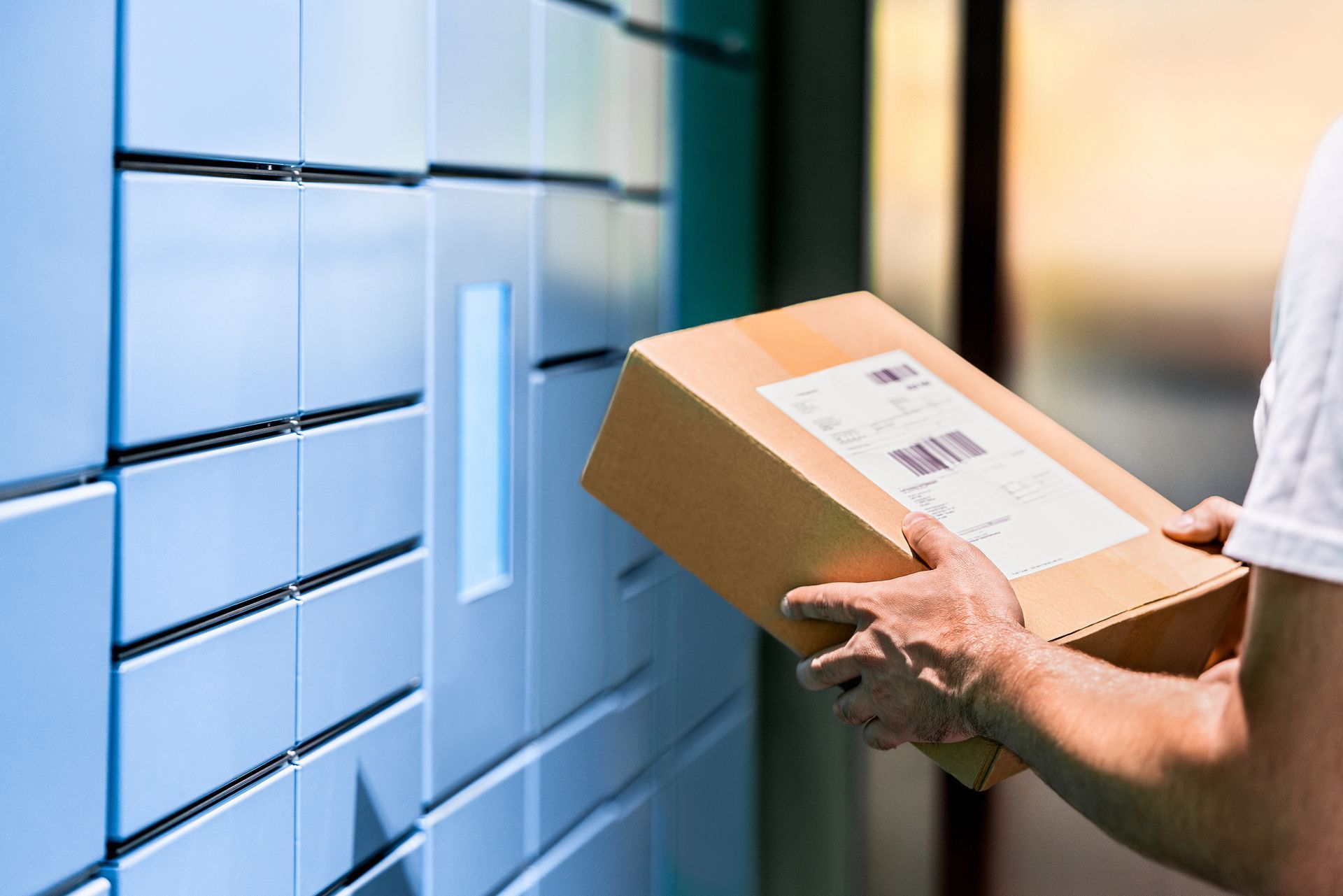 Person carrying a cardboard package beside blue lockers and a doorway