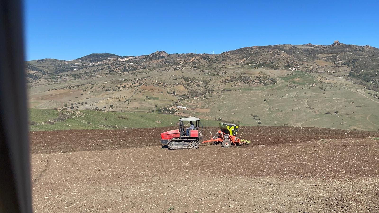 Un trattore rosso con cingoli traina attrezzature agricole attraverso un campo arato in una zona montuosa.