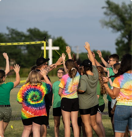 Group in colorful tie-dye shirts with raised hands outdoors near a volleyball court at sunset