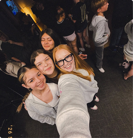 Four friends smiling in a crowded indoor selfie, one holding the camera overhead.