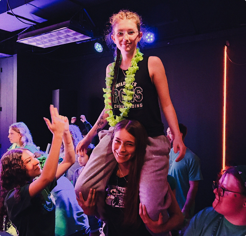 Person on someone’s shoulders in a neon-lit crowd, smiling and wearing a green lei at a lively event