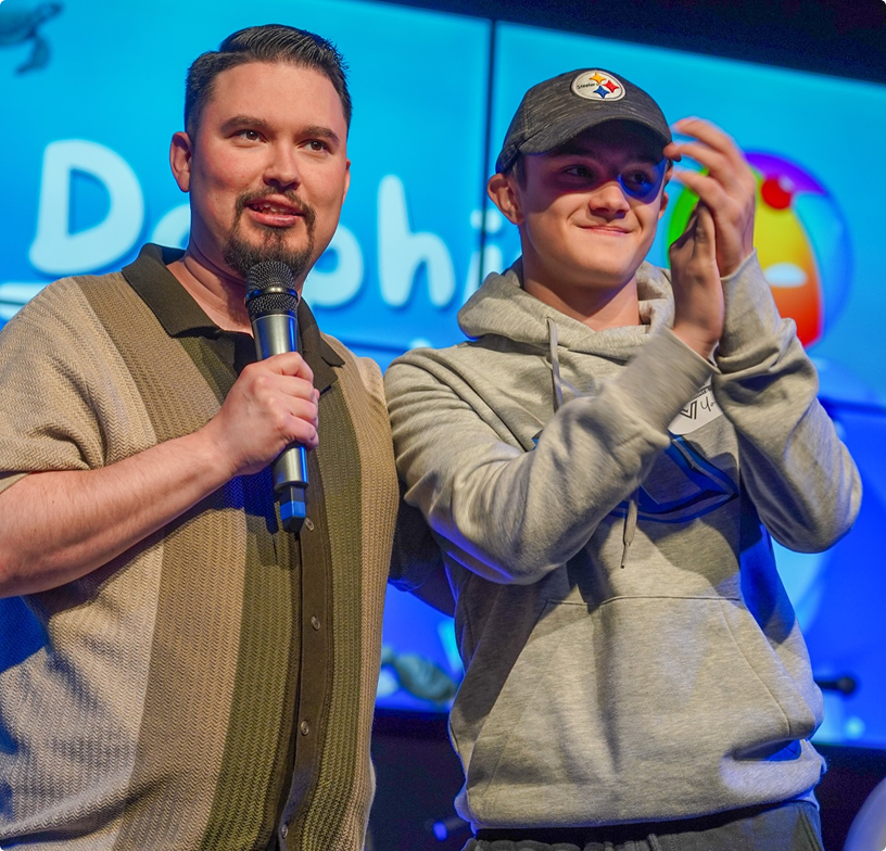 Two men onstage, one holding a microphone and the other clapping, before a blue-lit screen