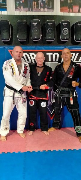 Three bald men in martial arts uniforms stand on a blue and pink mat.