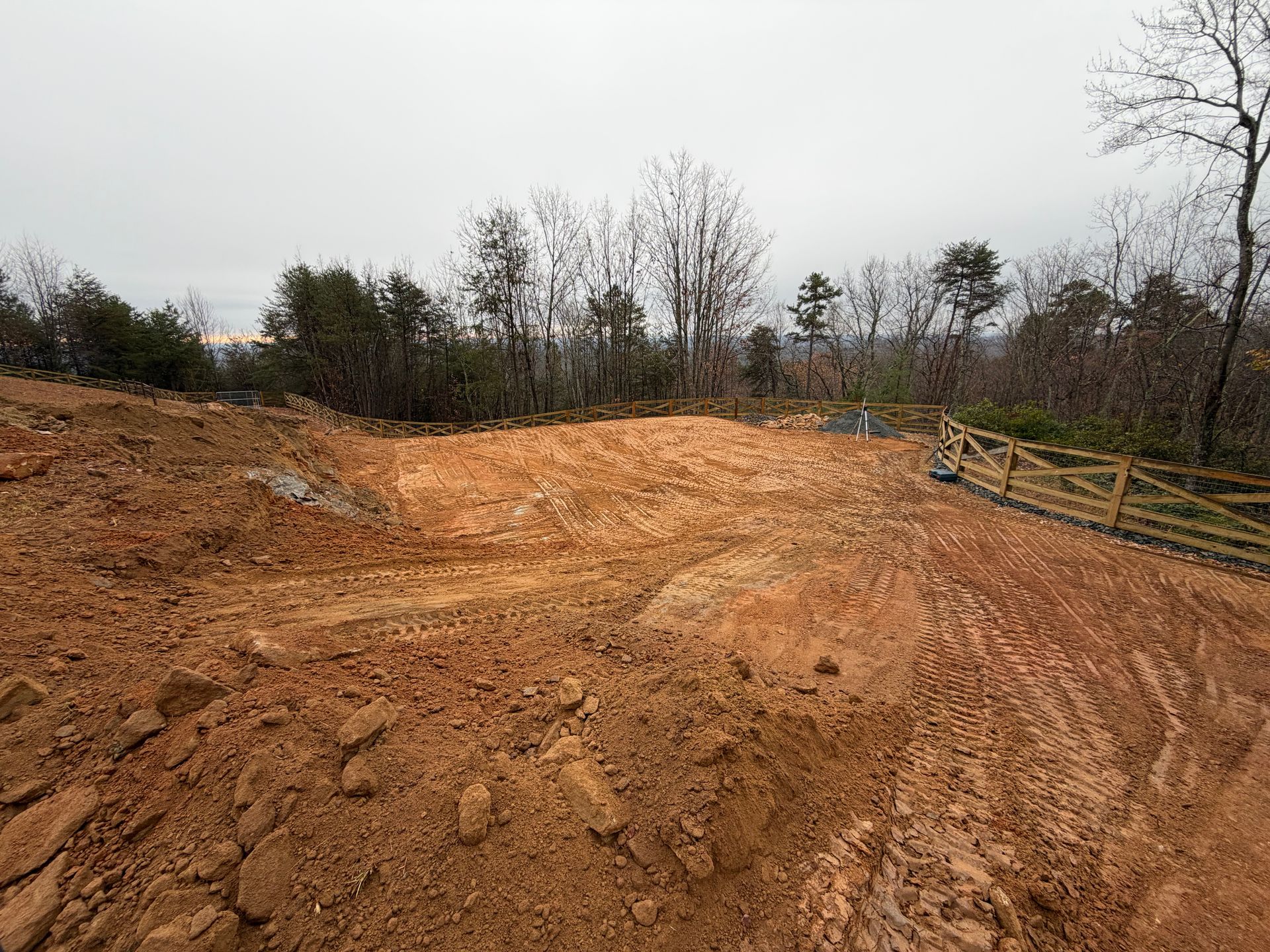 A graded construction site with exposed red dirt, a wooden fence on the right, and trees in the background under a gray sky.