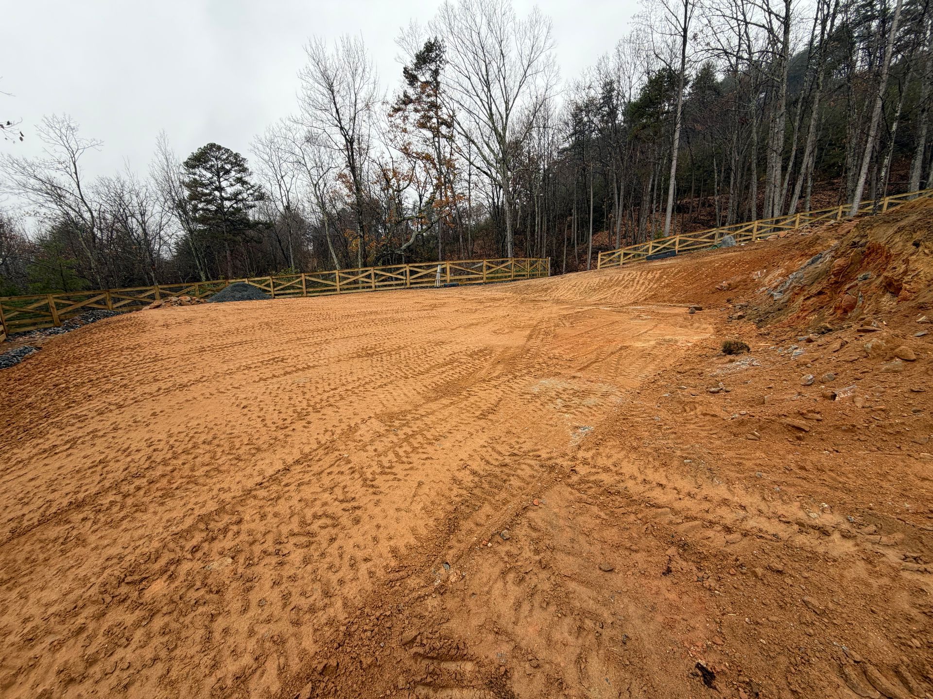A wide, cleared plot of brown, freshly graded dirt with tire tracks, set before a forested hillside and a wooden fence.