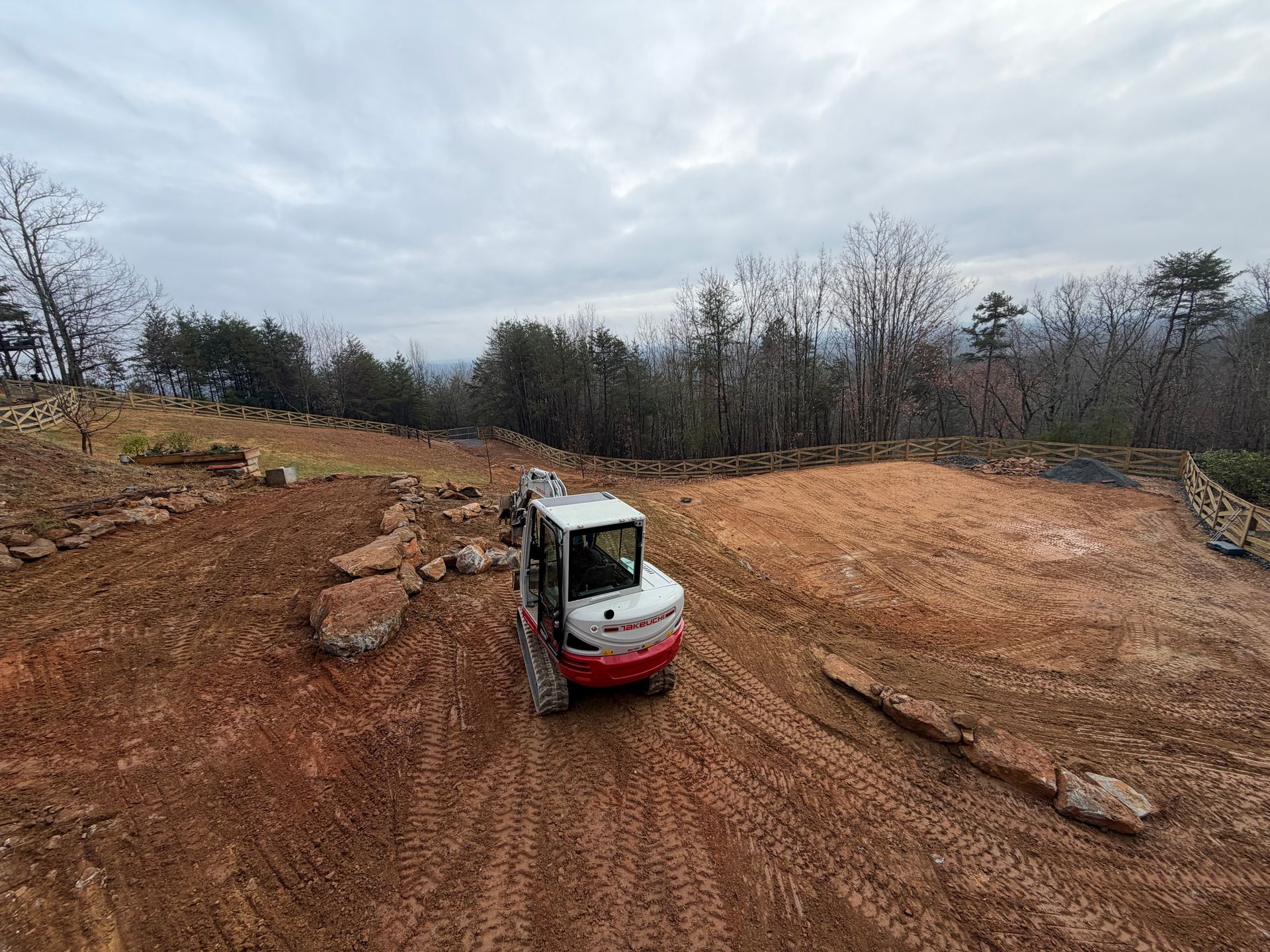 A white and red excavator sits on a dirt hillside under a cloudy sky, with trees in the background.