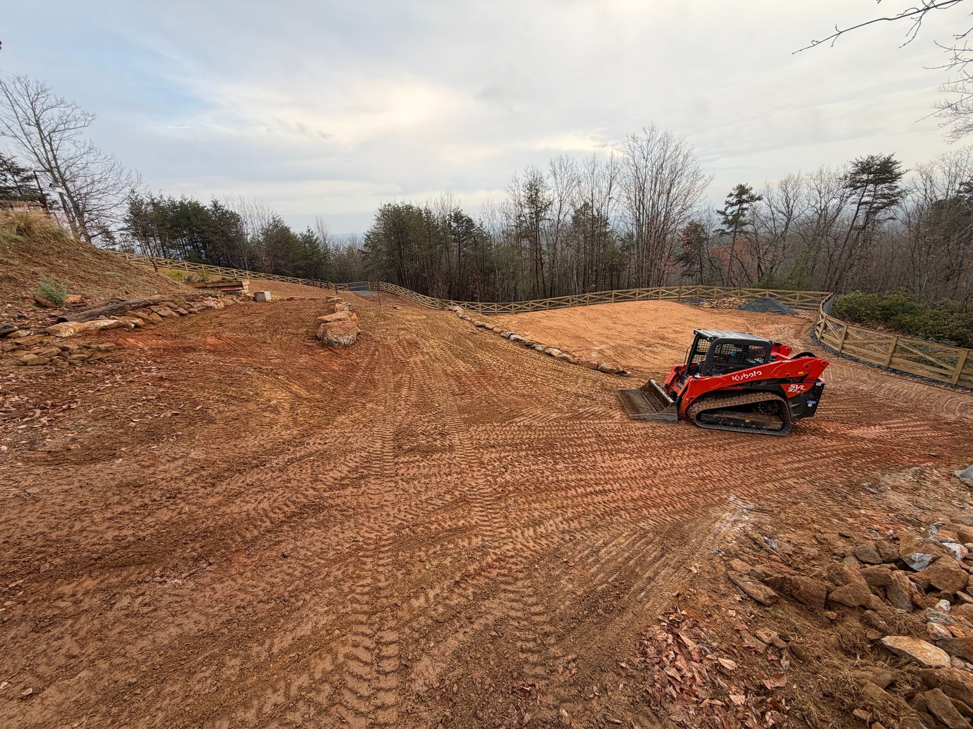 A red tracked skid steer loader works on a sloped, cleared dirt construction site under a cloudy sky.
