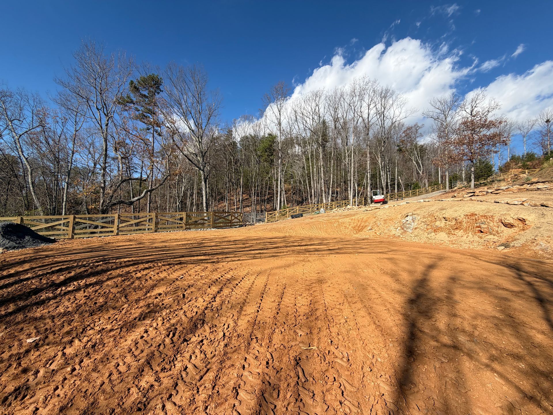 A graded, bare patch of red clay earth sits before a wooden fence and a forest of leafless trees under a bright blue sky.